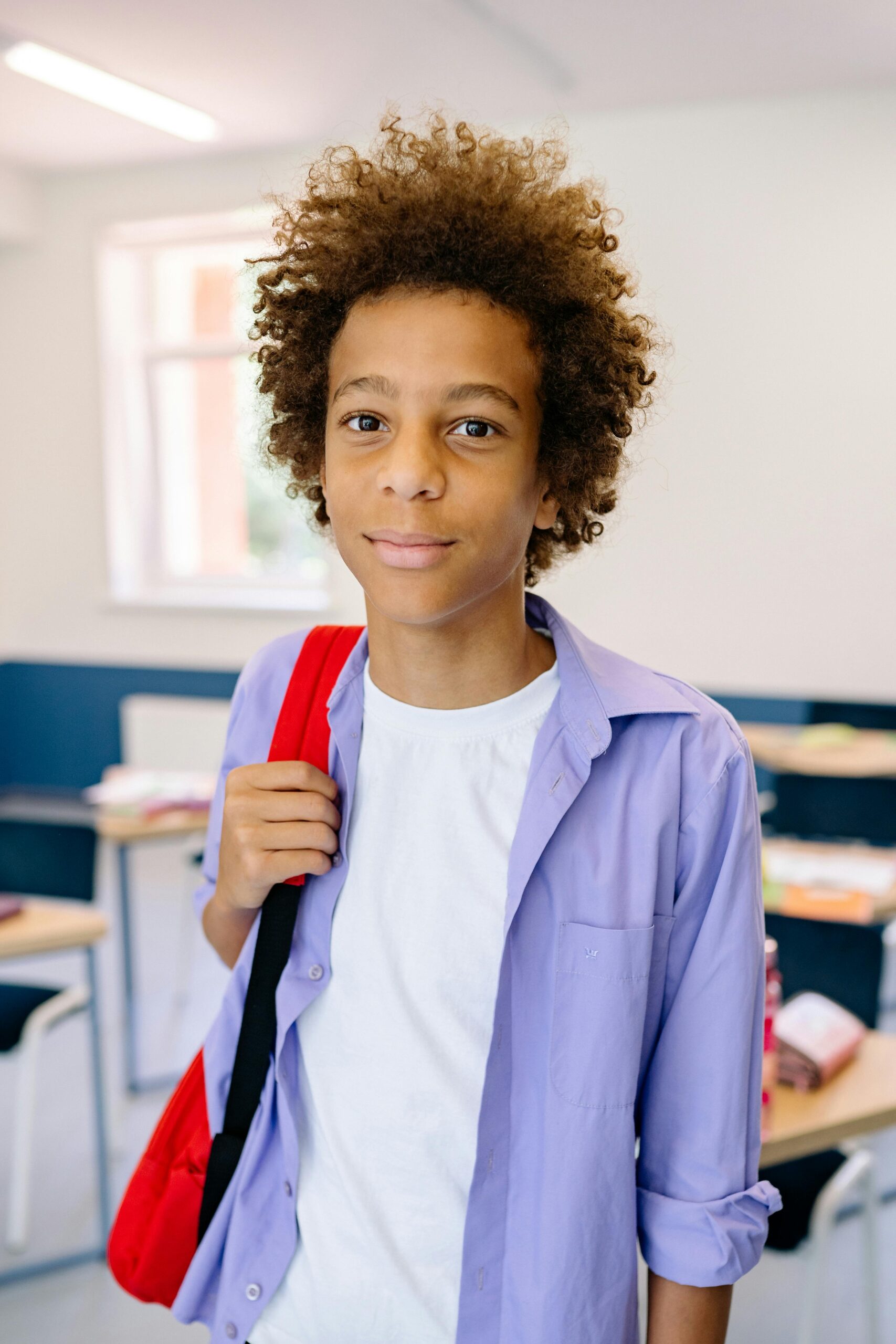 Happy adolescent boy with afro hair and backpack standing in a classroom setting.